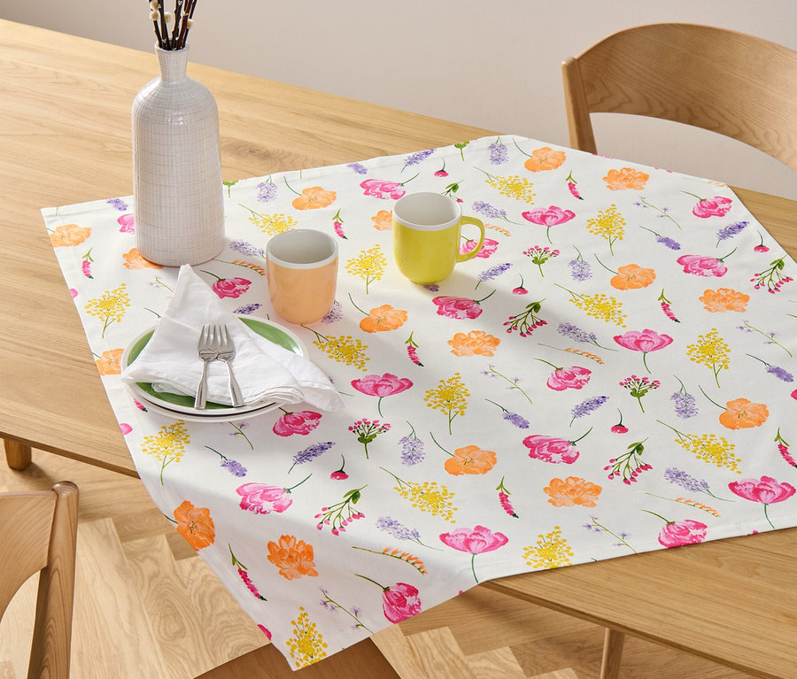 Sur une table en bois, une nappe en coton à motifs floraux est posée à côté d'un vase avec des brindilles et de deux tasses.