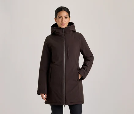 Femme aux cheveux foncés attachés en arrière, debout de face, portant une veste à capuche marron foncé avec les mains dans les poches.