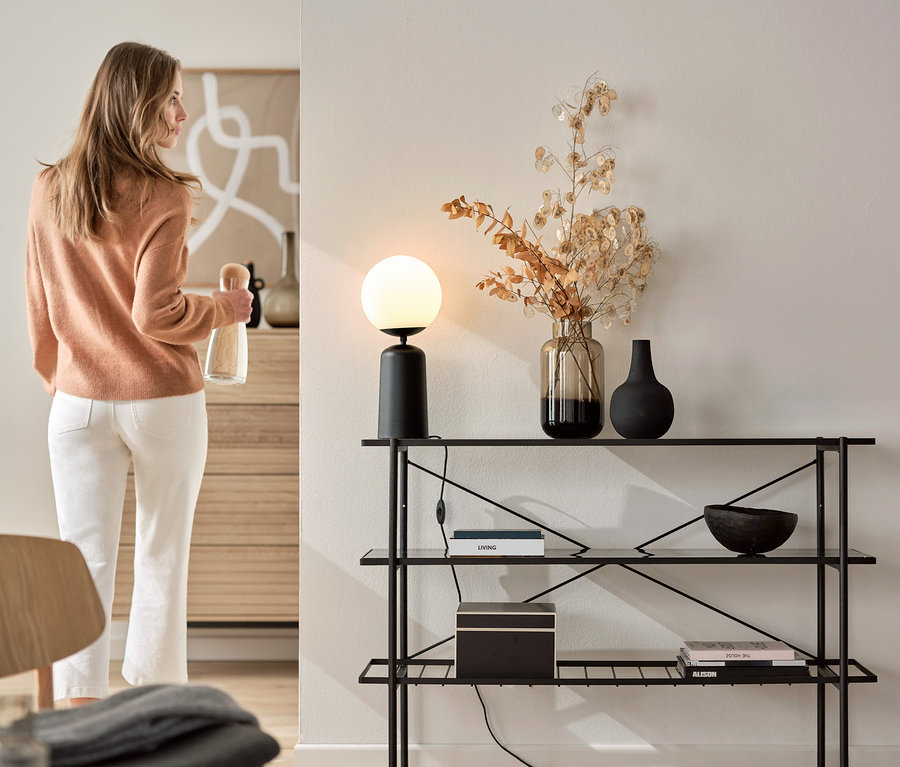 Femme tenant une bouteille à côté d'une console (largeur env. 120 cm) avec lampe de table noire avec globe en verre.