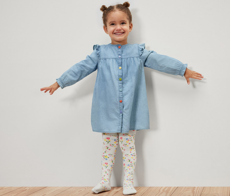 Une fillette souriante en robe en jean se tient debout, les bras tendus, et porte 5 collants pour enfant, floral.