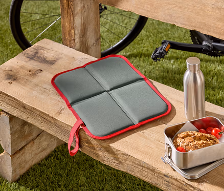 Un coussin de siège pliable gris avec un bord rouge est posé sur un banc en bois, à côté d'une boîte à pain et d'une bouteille en acier inoxydable. Un vélo est visible à l'arrière-plan.