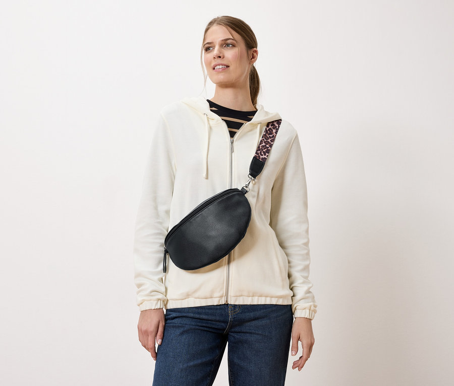 Une femme porte un sac banane noir avec une bandoulière à motif léopard sur une veste crème et un jean.