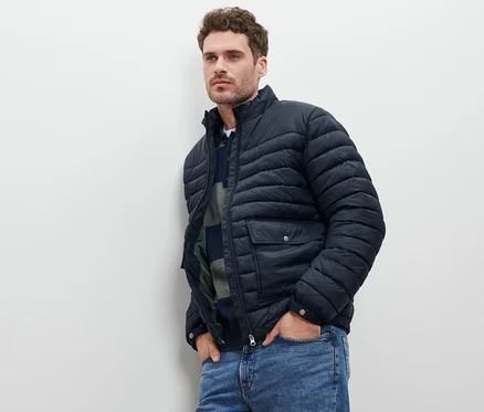 Un homme appuyé contre un mur portant une veste matelassée bleu marine et un jean.