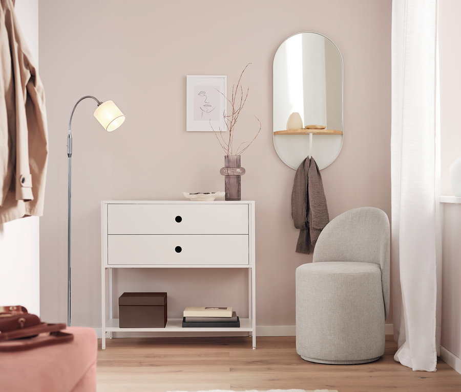 Une commode blanche avec deux tiroirs est placée à côté d'un miroir et d'un tabouret gris dans une pièce.