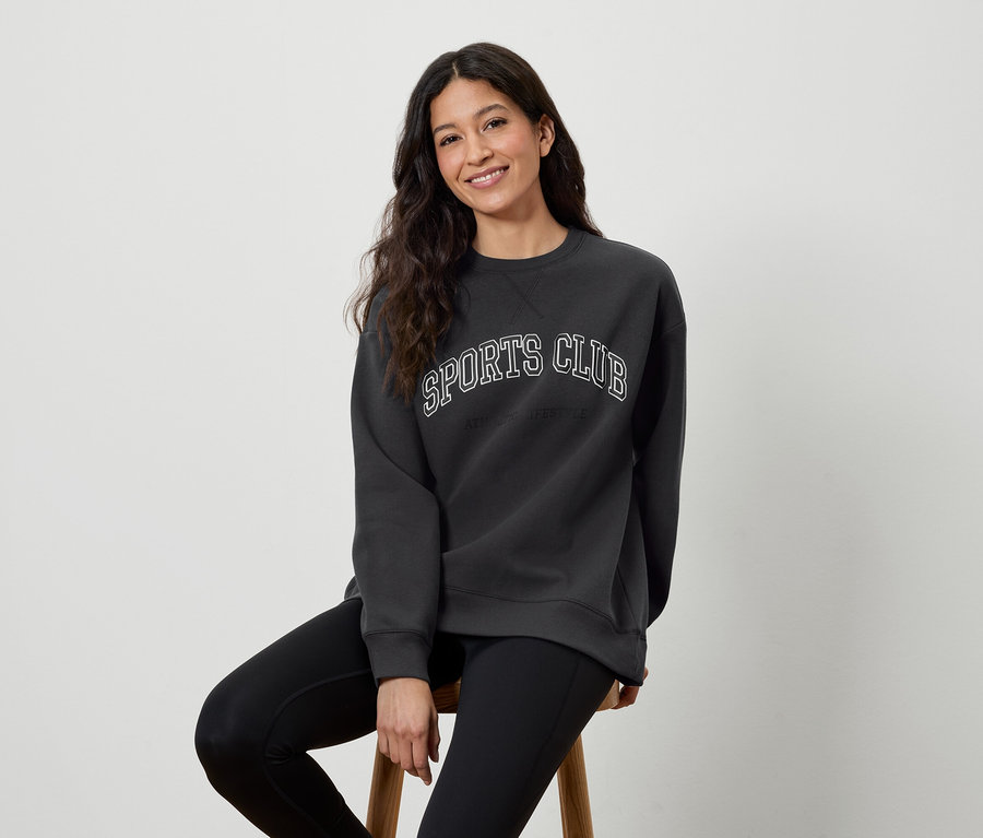 Une femme est assise sur un tabouret et porte un sweat-shirt gris foncé avec l'inscription SPORTS CLUB et des leggings noirs.