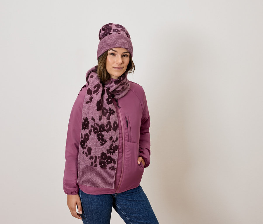Une femme porte une veste rose, un jean, une écharpe et un bonnet à fleurs.