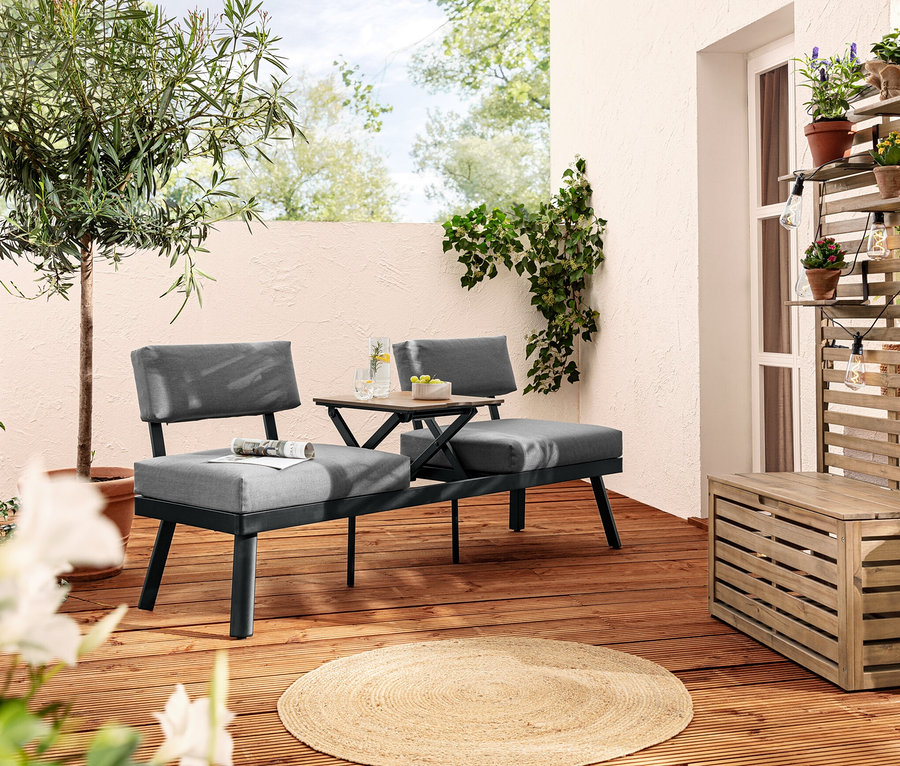 Deux canapés-lits 2 en 1 gris avec table centrale relevable sont posés sur une terrasse en bois. Une guirlande lumineuse à LED pour l’extérieur est accrochée à une structure en bois avec des fleurs.