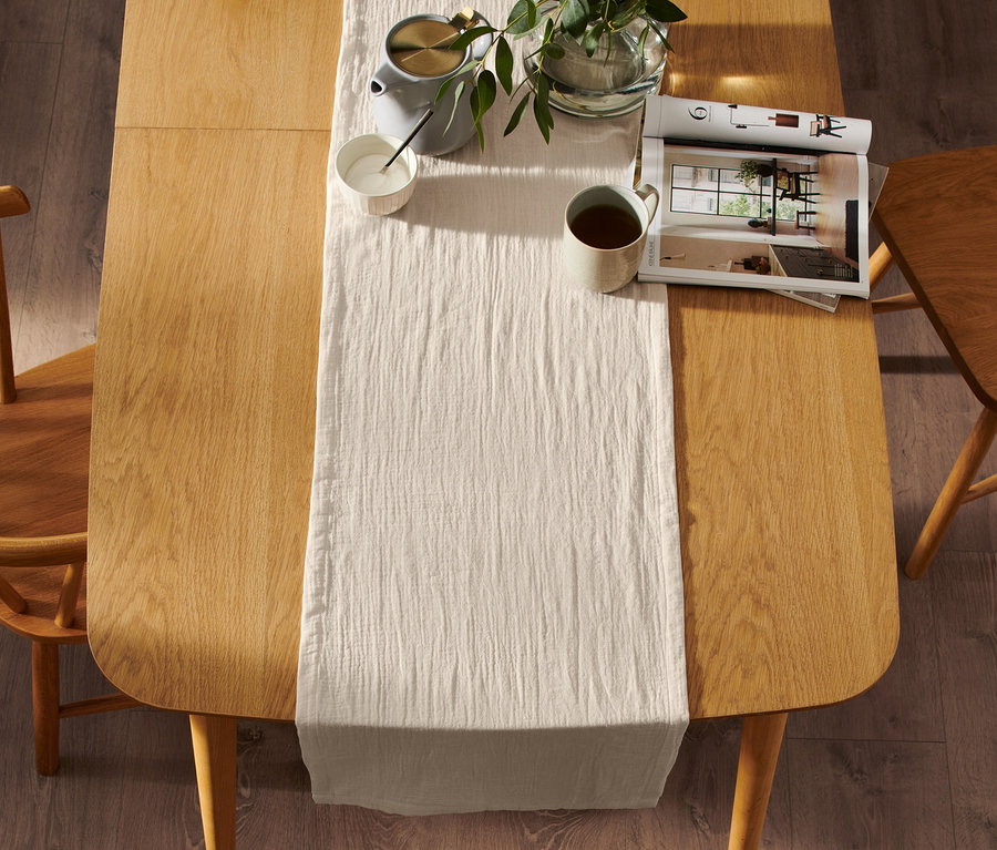 Un chemin de table beige en mousseline est disposé sur une table avec une tasse, une théière et une plante.