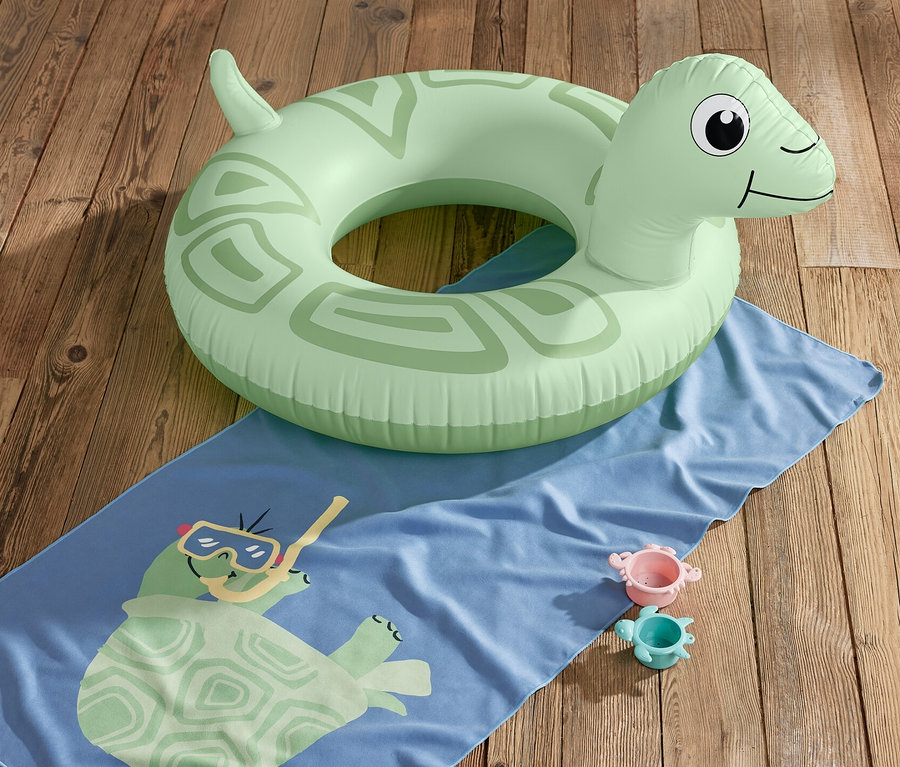 Une bouée « tortue » verte est posée sur une serviette bleue avec un motif de tortue et deux jouets sur un plancher en bois.
