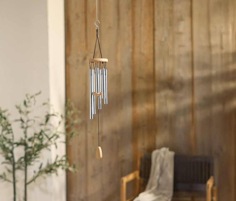 Un carillon éolien avec des tubes argentés est suspendu devant un mur en bois, à côté d'une plante et d'une chaise.