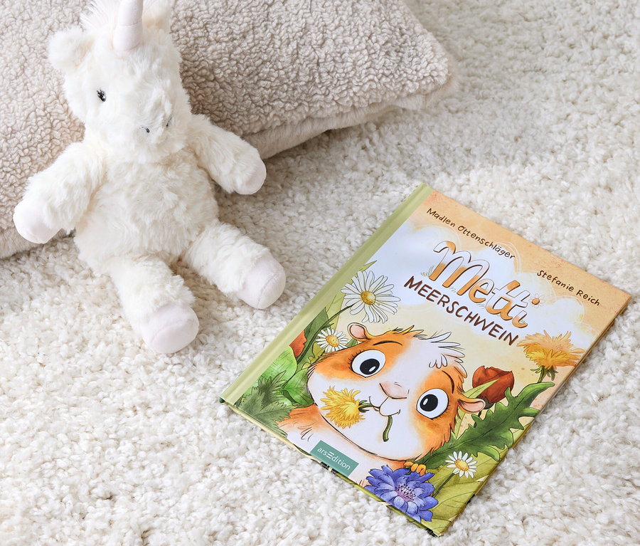 Une peluche licorne est allongée à côté du livre « Metti, le cochon d’Inde » sur un tapis blanc.