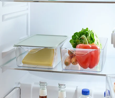 Dans le réfrigérateur, il y a des aliments sur les étagères : une pomme et un citron dans des récipients ronds, du fromage dans une boîte en plastique avec un couvercle, des légumes (salade, poivron, carottes) dans une boîte en plastique et des bouteilles de boissons dans un support.