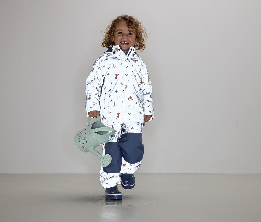 Un enfant aux cheveux bouclés portant une veste et un pantalon avec un imprimé animal tient un arrosoir vert.