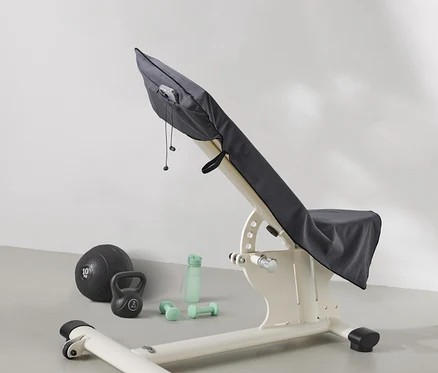 Un banc de musculation est recouvert d'un tissu gris. À côté du banc se trouvent un ballon de médecine, une kettlebell, une bouteille d'eau et des haltères.