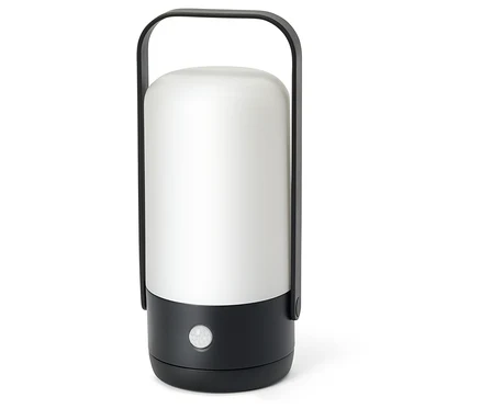 Lampe portable cylindrique avec un dessus blanc translucide et une base noire. Elle possède une poignée arquée noire et un petit capteur blanc sur le devant de la base.