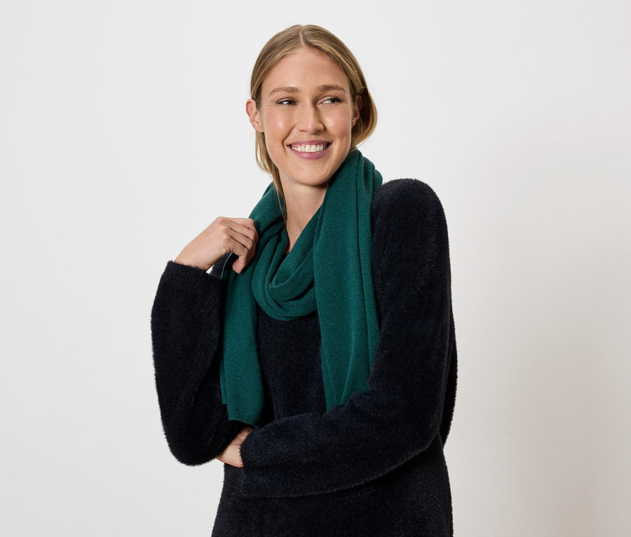 Une femme blonde souriante pose sur un fond clair avec une écharpe vert foncé et un pull.