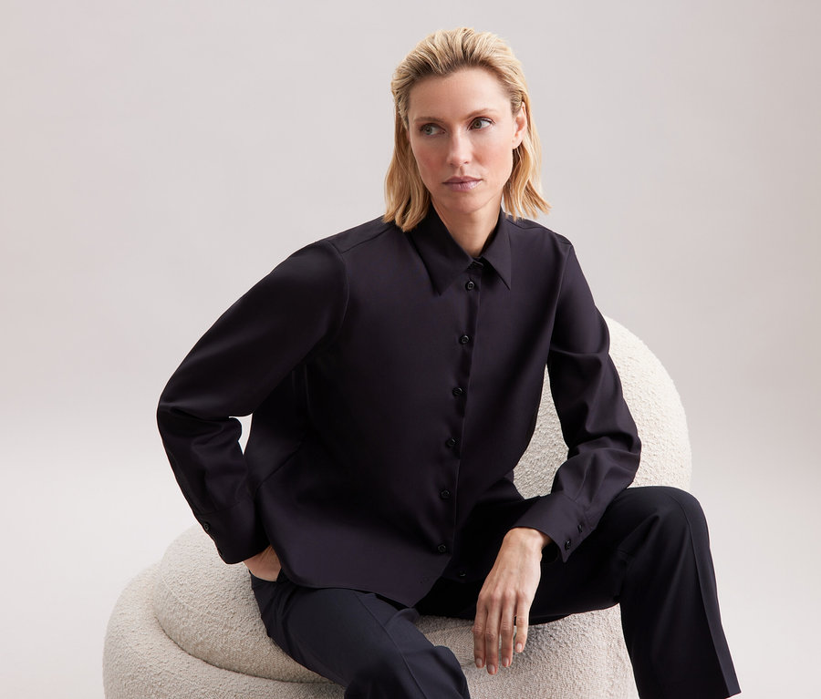 Une femme blonde est assise sur un fauteuil beige et porte une chemise noire et un pantalon.