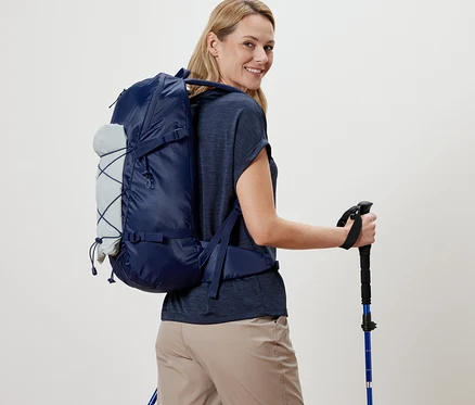 Femme avec un sac à dos d’extérieur et des bâtons de randonnée regardant par-dessus son épaule.