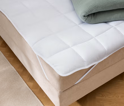 Un protège-matelas blanc matelassé avec des élastiques aux coins est posé sur un lit beige. Une couverture verte pliée est posée sur le dessus.