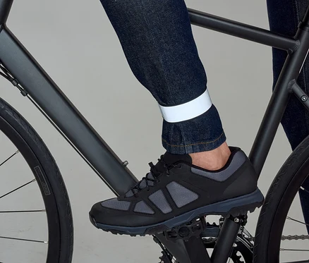 Une jambe en jean foncé avec une bande réfléchissante à la cheville pédale un vélo noir avec une chaussure noire et grise.