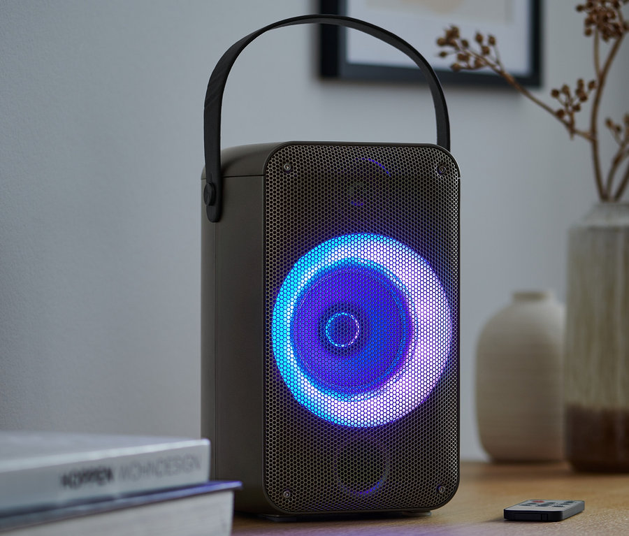 Un haut-parleur de fête Bluetooth® avec effets de lumière est posé sur une table à côté de livres et d'une télécommande.
