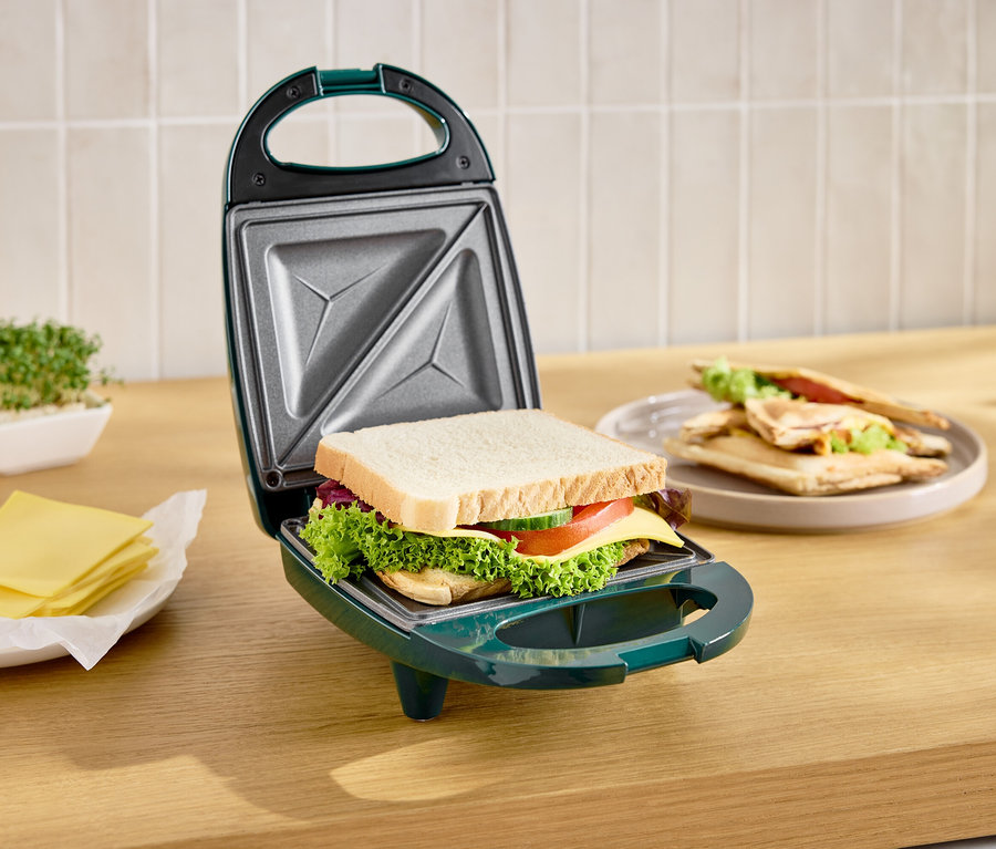 Appareil à sandwich vert ouvert avec un toast à l'intérieur, à côté une assiette avec des toasts finis et du fromage.