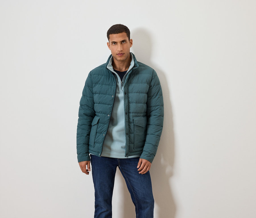 Un homme pose sur un fond blanc vêtu d'une veste bleue, d'un pull bleu et d'un jean.