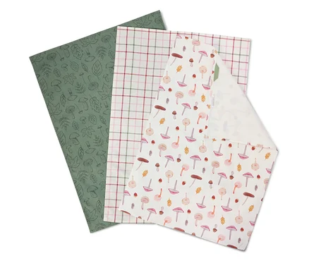 Trois serviettes en tissu superposées : une vert olive à motifs de feuilles, une à carreaux et une blanche à motifs de champignons.