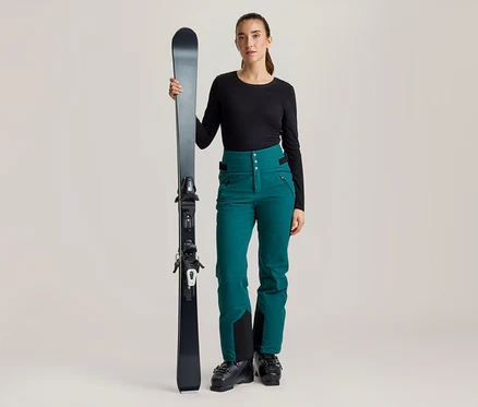 Une femme tient un ski noir. Elle porte un haut noir à manches longues, un pantalon de ski sarcelle et des chaussures de ski noires.