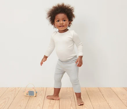 Un enfant debout sur un plancher en bois porte 3 bodys côtelés manches longues pour bébé et un legging gris.