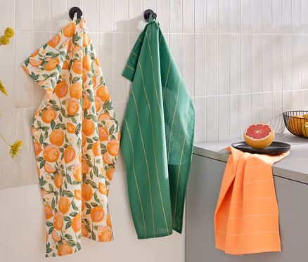 Trois torchons sont accrochés au mur : un avec un imprimé d'agrumes, un vert rayé et un orange. Un demi-pamplemousse et un bol de fruits sont posés sur le plan de travail.
