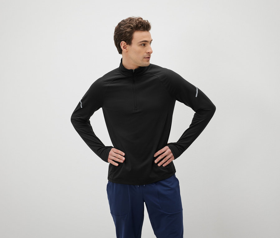 Un homme aux cheveux bruns pose les mains sur les hanches, vêtu d'un t-shirt noir à manches longues et d'un pantalon bleu.