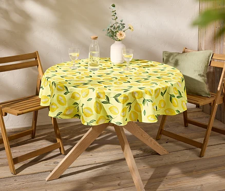 Une table ronde avec une nappe jaune à motif de citron est placée entre deux chaises en bois.
