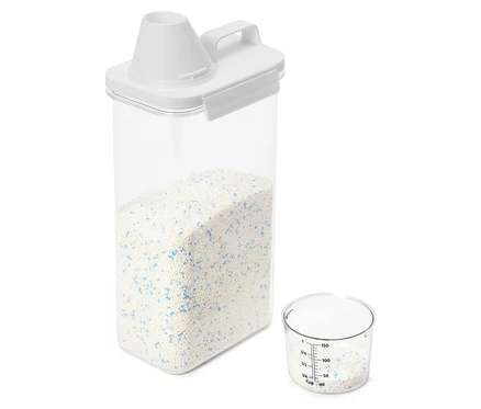 Un récipient transparent avec un couvercle blanc est rempli de lessive. À côté se trouve un gobelet doseur, qui contient également de la lessive.