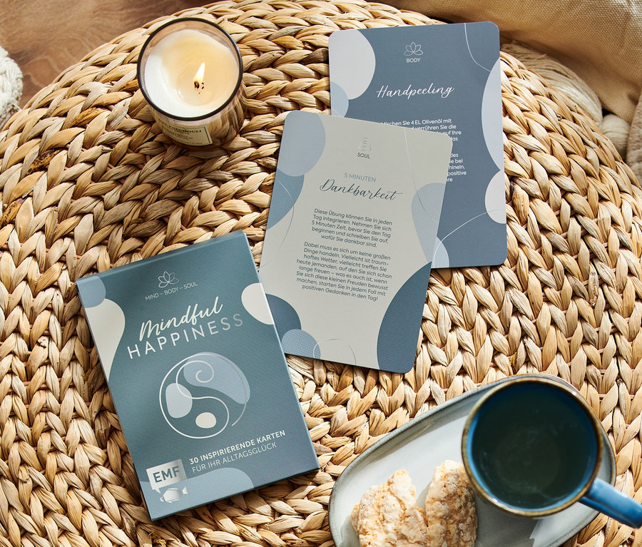 Sur un tabouret en rotin sont posés une bougie, une tasse, des biscuits et la boîte de cartes « Mindful Happiness ».