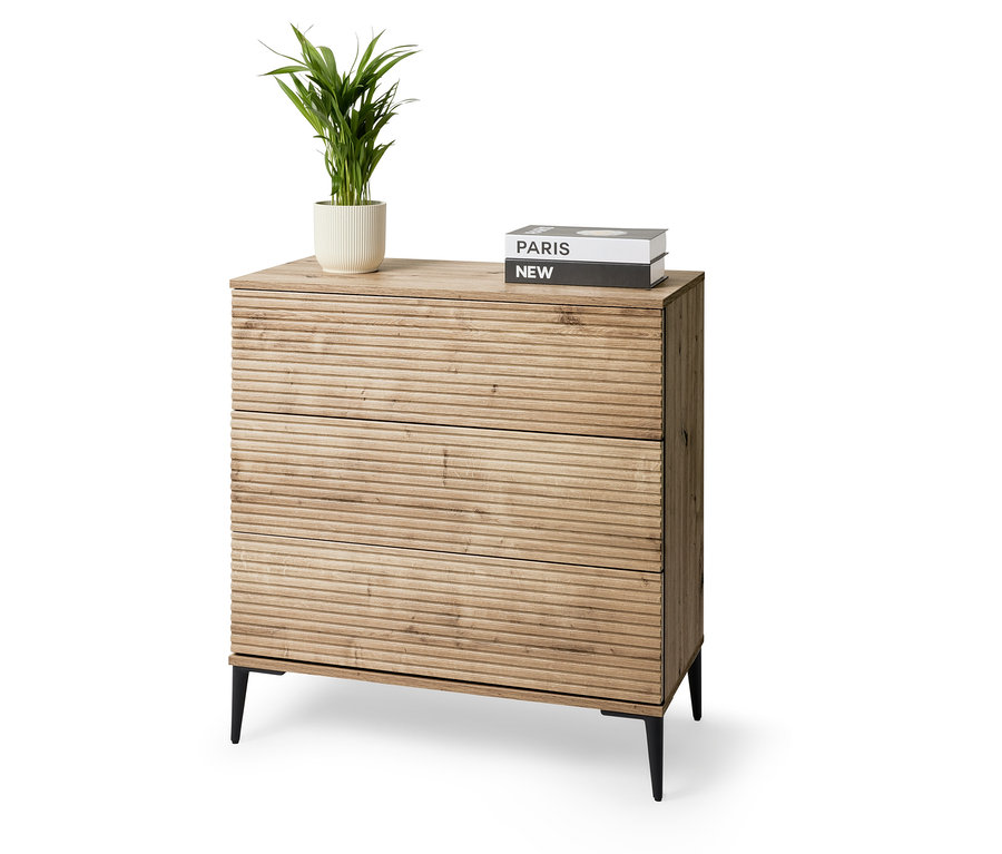 Une commode à trois tiroirs se dresse sur un fond blanc. Sur la commode, il y a une plante en pot et deux livres.