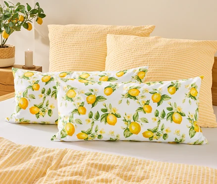 Sur un lit, deux coussins à motifs de citrons sont placés devant deux coussins jaunes. À gauche, un citronnier est posé à côté d'une bougie.