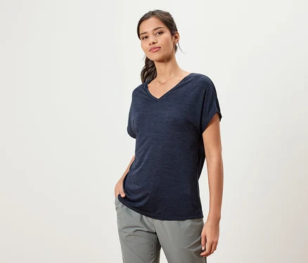 Une femme portant un t-shirt foncé et un pantalon gris se tient devant un fond blanc.