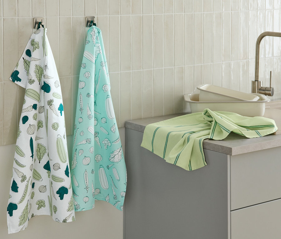 Deux linges de cuisine à motifs de légumes sont accrochés au mur, un autre linge de cuisine vert est posé sur le plan de travail.