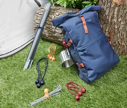 Sur l'herbe se trouvent une tente, des piquets de tente attachés par 12 élastiques, et un sac à dos bleu.