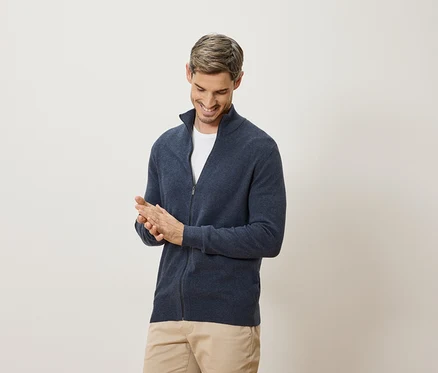 Un homme sourit et se frotte les mains. Il porte un cardigan bleu marine et un pantalon beige.