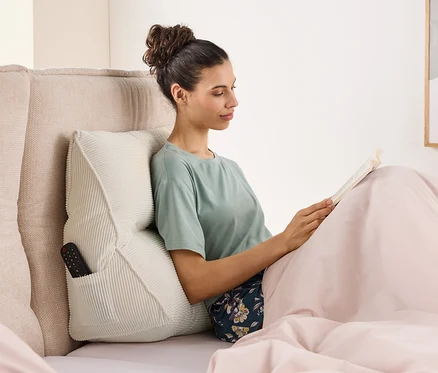 Une femme est assise dans son lit en train de lire un livre, adossée à un coussin avec une télécommande dans la poche latérale.