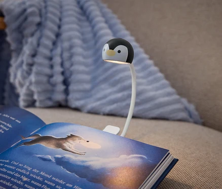 Un livre ouvert avec une couverture bleu foncé repose sur un canapé beige, éclairé par une lampe à pince. La lampe a un bras flexible blanc et une tête en forme de pingouin noir et blanc, projetant de la lumière sur les pages du livre. La page de droite présente une illustration d'un loup bondissant à travers un ciel nuageux et bleu. Une couverture texturée bleu clair est drapée sur le dossier du canapé en arrière-plan.