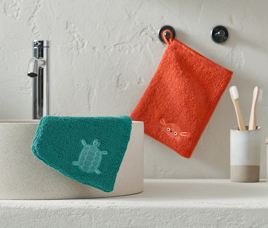 Un gant de toilette vert avec une tortue repose sur un lavabo, un gant de toilette orange avec un crabe est accroché au mur.