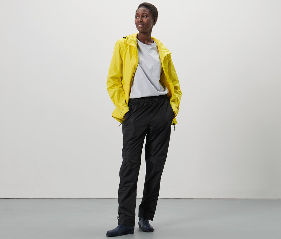 Une femme noire se tient debout, les mains dans les poches d'un pantalon de pluie high-tech noir et d'une veste de pluie jaune.