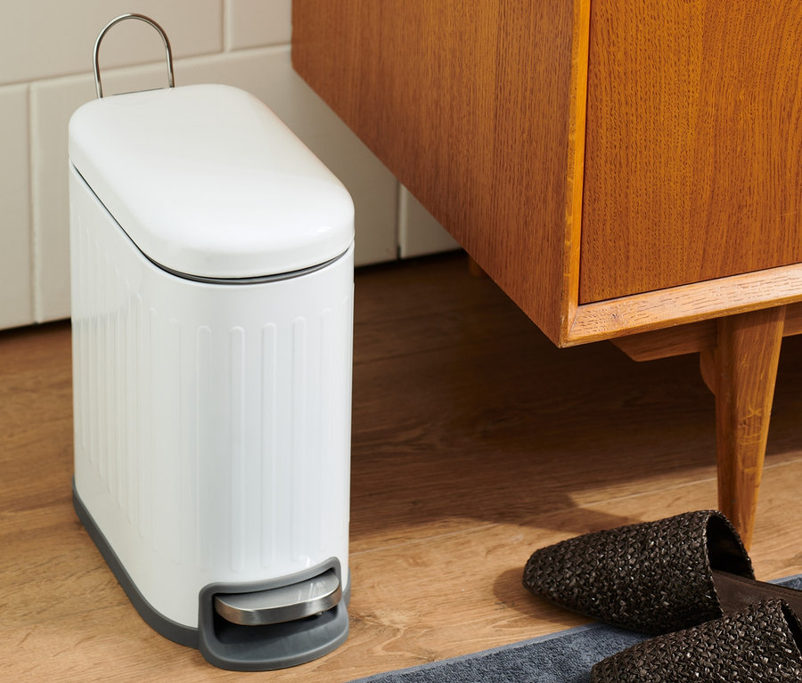 Dans une salle de bain, une poubelle blanche est posée à côté d'un meuble en bois marron et de chaussons sur un tapis gris.