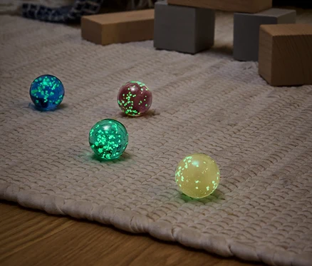 Quatre boules lumineuses de différentes couleurs (bleu, rouge, vert et jaune) sur un tapis beige avec des jouets en bois en arrière-plan.