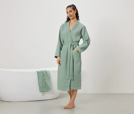 Une femme se tient pieds nus dans un peignoir vert menthe avec une ceinture et des poches, une main dans la poche. Une baignoire avec une serviette vert menthe est visible en arrière-plan.