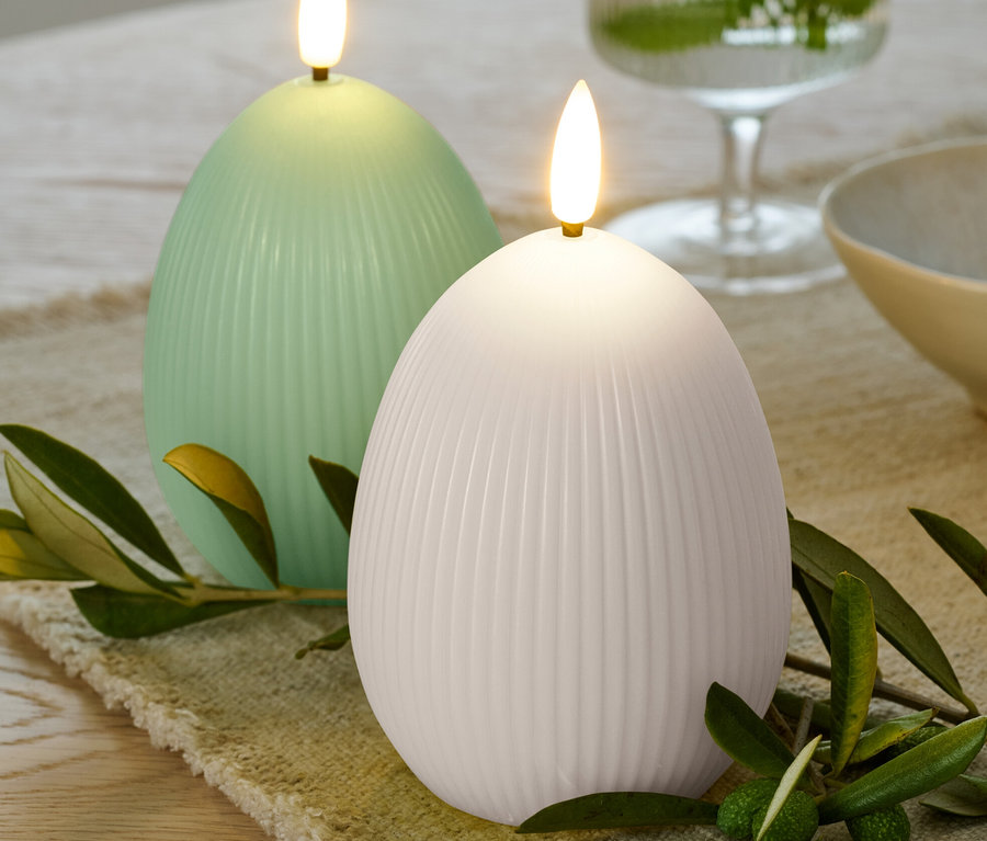 Deux bougies à LED en cire véritable en forme d'œuf, une blanche et une vert clair, posées sur un chemin de table beige avec des branches d'olivier vertes.