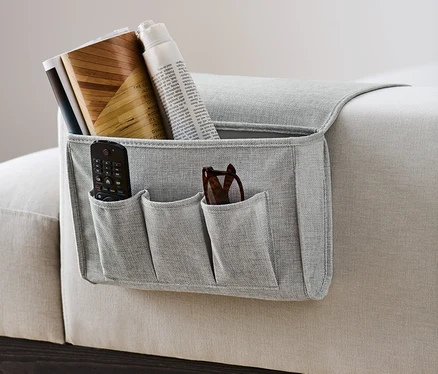 Un rangement de lit ou de canapé gris est accroché sur le côté d'un canapé et contient une télécommande, des lunettes, des livres et un journal.